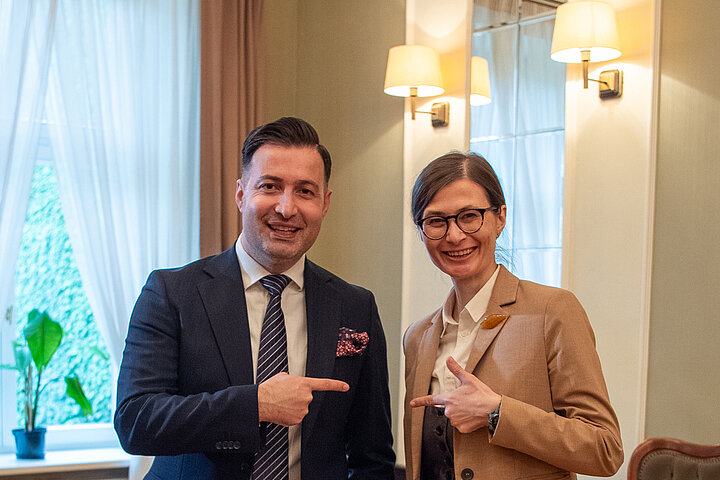 A man and a woman stand side by side, smiling at the observer. They have their index fingers extended, pointing at each other.