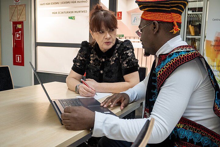 A woman and a man are talking at a desk. The man is wearing an exotic, colorful hat.