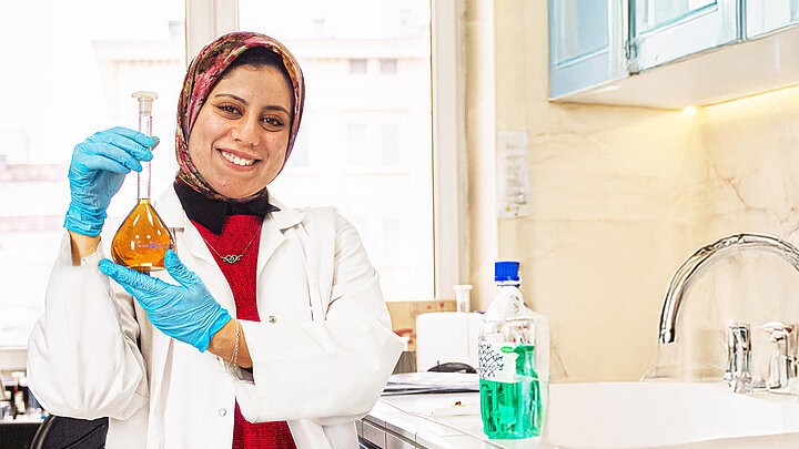 Safya Elsharkawy in a laboratory, holding a flask with an orange liquid and smiling.