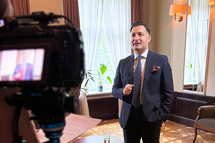 A man in a navy blue suit stands in a classically decorated room. In front of him is a camera on a tripod.