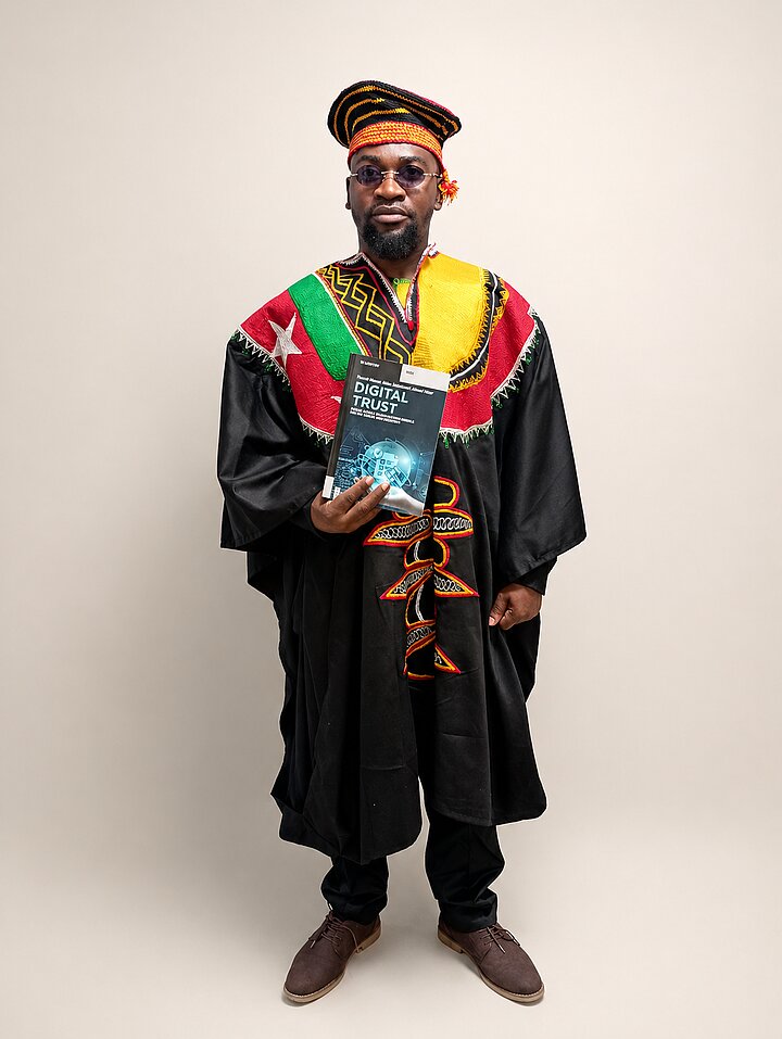 A man in an African ethnic tunic poses for a photo. He holds a book titled Digital Trust.