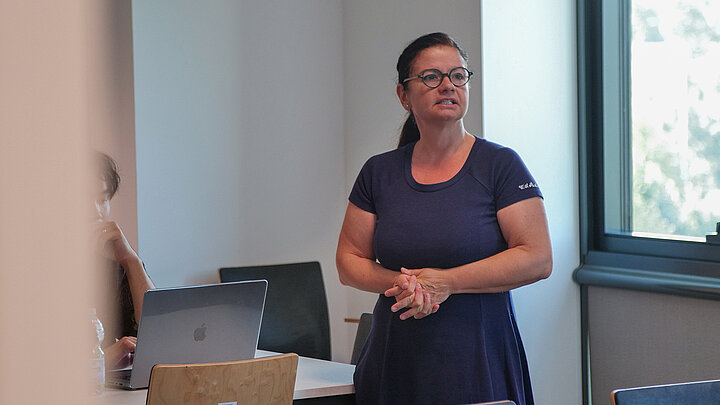 A woman stands in a classroom. A student sits at a desk.